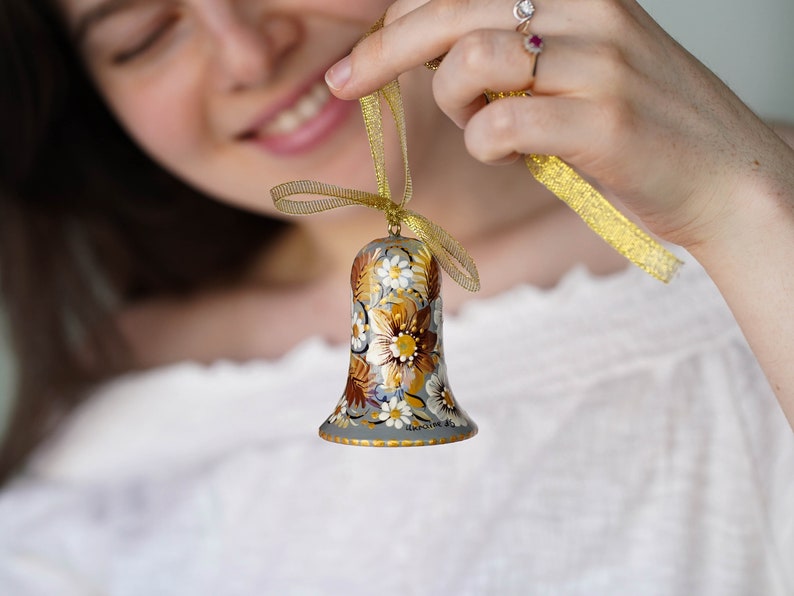Op de afbeelding: Een grijze bel ornament met een gouden lint en bloemenpatroon. De bel is geschilderd met witte, gouden en bruine bloemen.