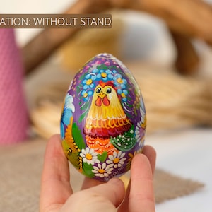 May include: A hand is holding a purple wooden egg painted with a colourful rooster and floral design. The egg is decorated with yellow, red, green, blue and white paint. The text "Ukraine" is visible on the egg.