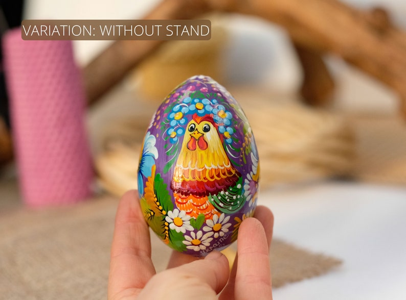 May include: A hand is holding a purple wooden egg painted with a colourful rooster and floral design. The egg is decorated with yellow, red, green, blue and white paint. The text "Ukraine" is visible on the egg.