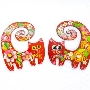 May include: Two red wooden cat figurines with painted floral designs. The cats have curled tails and are painted with bright colours, including pink, yellow, green, and white.