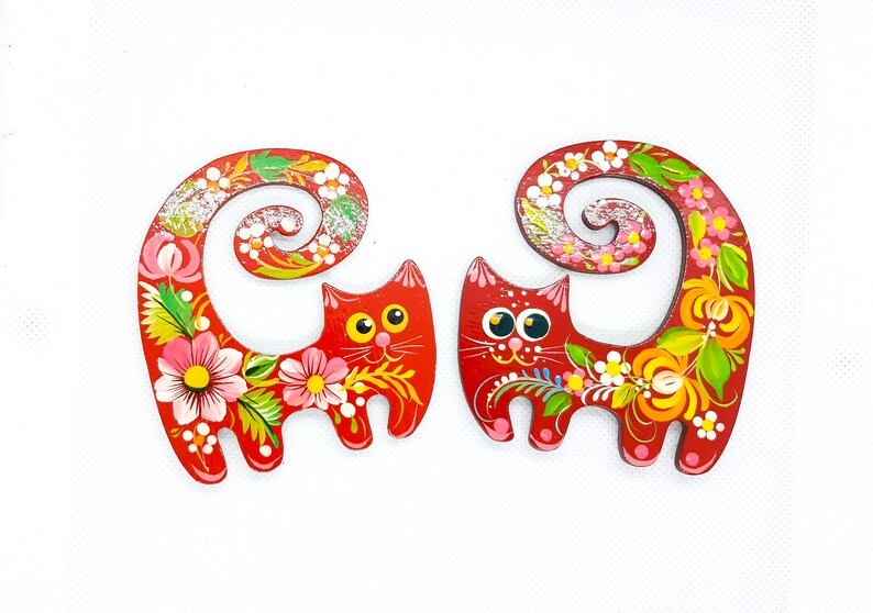 May include: Two red wooden cat figurines with painted floral designs. The cats have curled tails and are painted with bright colours, including pink, yellow, green, and white.