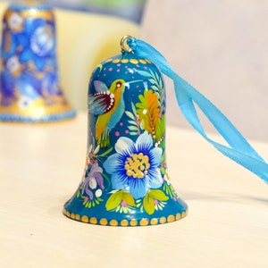 May include: A teal blue bell ornament with a painted hummingbird and floral design. The bell has a gold trim and is attached to a blue ribbon.