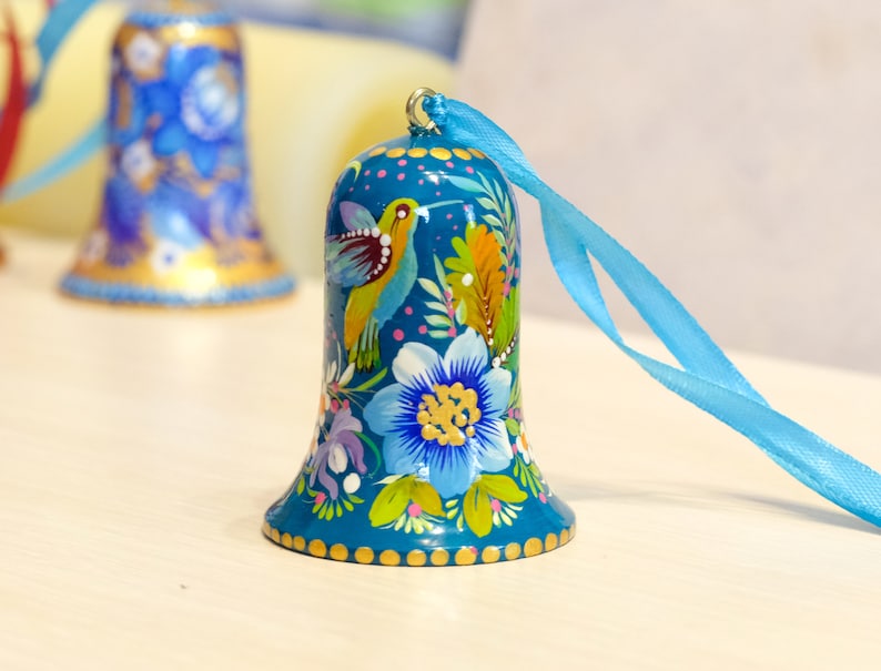 May include: A teal blue bell ornament with a painted hummingbird and floral design. The bell has a gold trim and is attached to a blue ribbon.
