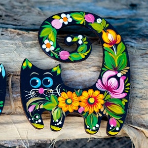 May include: Two black wooden cat figurines painted with colorful flowers and swirls. The cat on the right has its tail curled up in a circle.