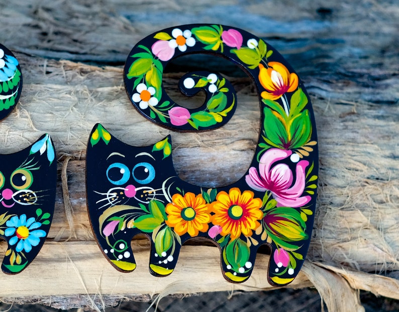 May include: Two black wooden cat figurines painted with colorful flowers and swirls. The cat on the right has its tail curled up in a circle.