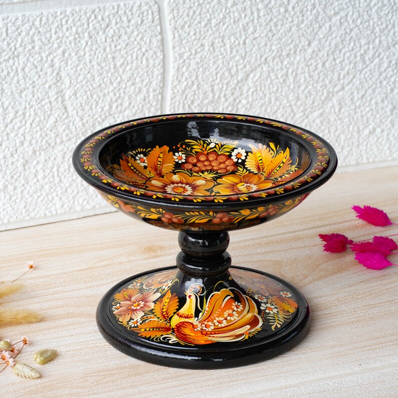 May include: A black wooden bowl with a pedestal base, hand-painted with a floral design in shades of orange, yellow, and red. The bowl features a stylized bird with a long tail.