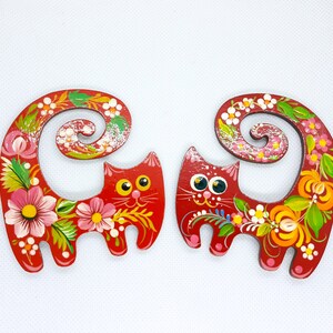 May include: Two red wooden cat figurines with curled tails. The cats are painted with colourful floral designs and have black eyes.