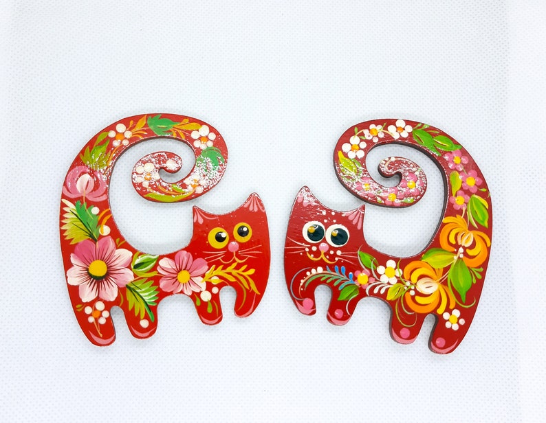 May include: Two red wooden cat figurines with curled tails. The cats are painted with colourful floral designs and have black eyes.