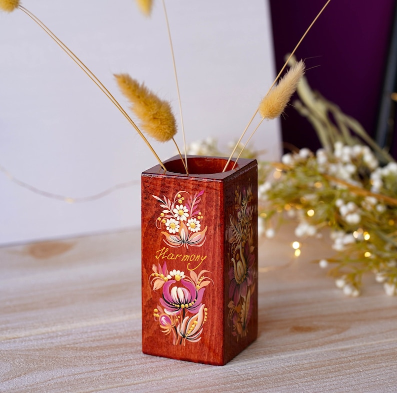 Puede incluir: Un jarr&oacute;n de madera con un dise&ntilde;o floral pintado a mano y la palabra "Armon&iacute;a" en letras doradas. El jarr&oacute;n est&aacute; pintado en tonos de rojo, rosa, blanco y dorado.