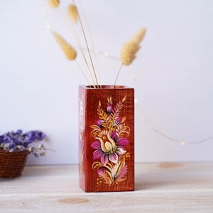 Puede incluir: Un jarr&oacute;n de madera cuadrado con un dise&ntilde;o floral pintado en la parte delantera. El jarr&oacute;n est&aacute; pintado en tonos de rojo, naranja, amarillo, rosa y morado. El jarr&oacute;n est&aacute; sobre una superficie de madera clara.