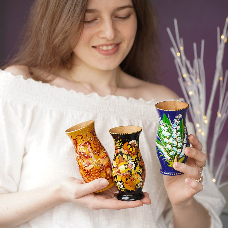 May include: Three hand-painted wooden vases with floral designs. The vases are blue, yellow, and black with white floral patterns.