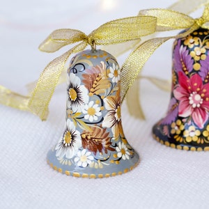 Op de afbeelding: Twee handgeschilderde kerstbelletjes met bloemmotieven. Het belletje links is grijs met witte en gouden bloemen. Het belletje rechts is zwart met roze, paarse en gouden bloemen. Beide belletjes hebben een gouden lint.