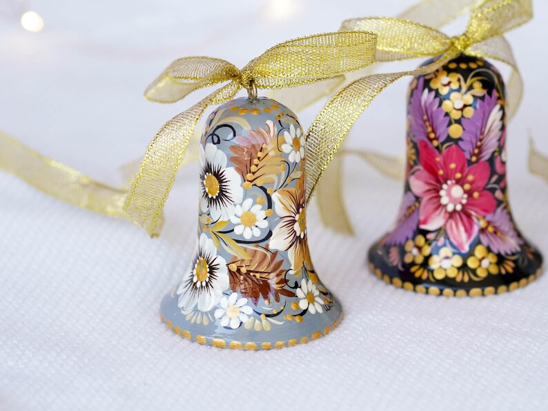 Op de afbeelding: Twee handgeschilderde kerstbelletjes met bloemmotieven. Het belletje links is grijs met witte en gouden bloemen. Het belletje rechts is zwart met roze, paarse en gouden bloemen. Beide belletjes hebben een gouden lint.