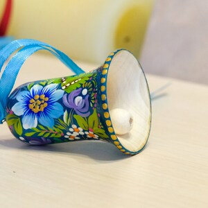 May include: A blue and green painted wooden bell with a white bead inside. The bell has a floral design with blue, yellow, and purple flowers. A blue ribbon is attached to the top of the bell.