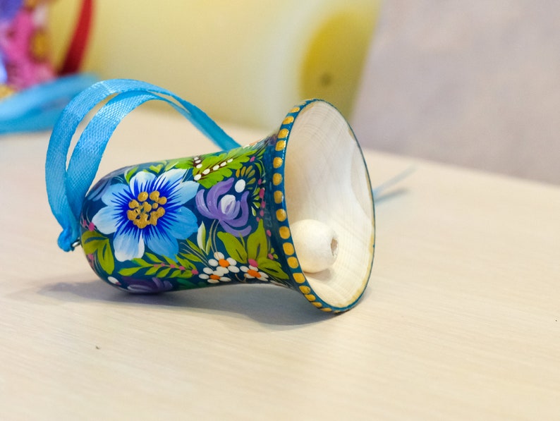 May include: A blue and green painted wooden bell with a white bead inside. The bell has a floral design with blue, yellow, and purple flowers. A blue ribbon is attached to the top of the bell.