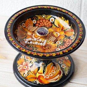May include: A black wooden trinket dish with a painted floral design and a gold bird on the base. The dish is filled with a watch and a pearl necklace.