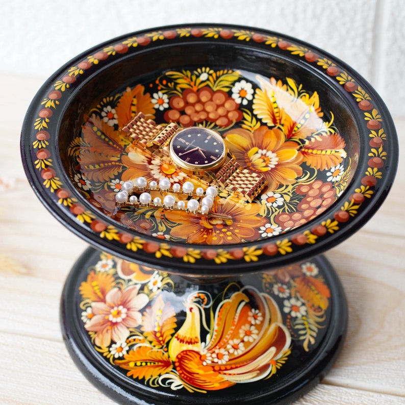 May include: A black wooden trinket dish with a painted floral design and a gold bird on the base. The dish is filled with a watch and a pearl necklace.