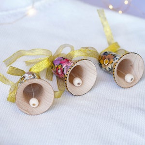 May include: Three wooden bells with floral patterns and gold ribbon. The bells are painted with pink, purple, yellow, and green flowers. The bells are hanging from the ribbon.