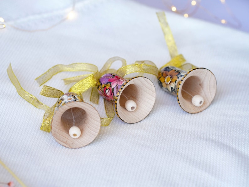 May include: Three wooden bells with floral patterns and gold ribbon. The bells are painted with pink, purple, yellow, and green flowers. The bells are hanging from the ribbon.