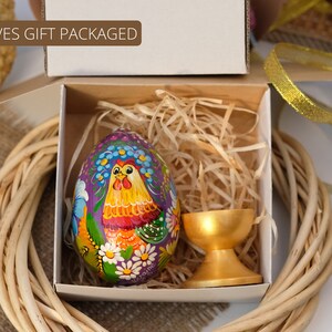 May include: A hand-painted wooden Easter egg featuring a rooster design in shades of purple, yellow, red, and green. The egg is presented in a white gift box with shredded paper and a gold egg stand.