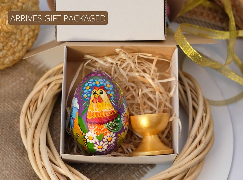 May include: A hand-painted wooden Easter egg featuring a rooster design in shades of purple, yellow, red, and green. The egg is presented in a white gift box with shredded paper and a gold egg stand.
