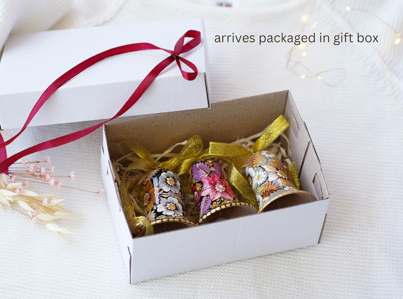 May include: Three hand-painted bells with floral designs in a white gift box with a red ribbon. The bells are decorated with gold accents and have a rustic, vintage feel.  Arrives packaged in a gift box.