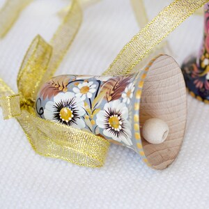 Op de afbeelding: Een handgeschilderde houten bel met een bloemenmotief in tinten wit, bruin en goud. De bel is vastgebonden met een gouden lint en heeft een kleine houten klopper.