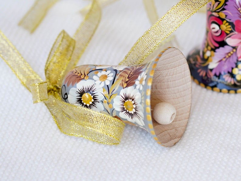 Op de afbeelding: Een handgeschilderde houten bel met een bloemenmotief in tinten wit, bruin en goud. De bel is vastgebonden met een gouden lint en heeft een kleine houten klopper.