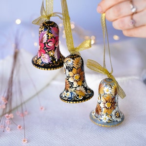 May include: Three hand-painted bells with floral designs. The bells are black, gold, and gray with intricate floral patterns. The bells are hanging from gold ribbon.