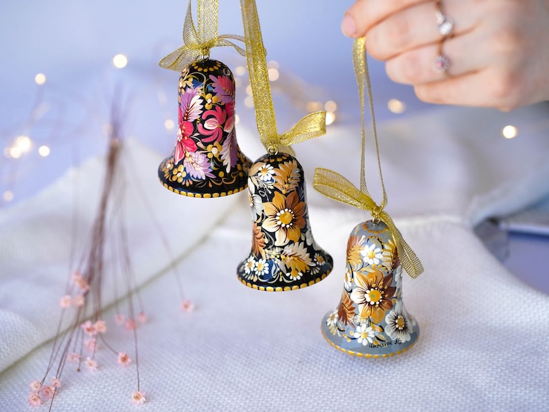 May include: Three hand-painted bells with floral designs. The bells are black, gold, and gray with intricate floral patterns. The bells are hanging from gold ribbon.