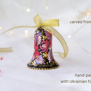 May include: A hand-painted wooden bell with a floral design in shades of pink, purple, and gold. The bell is decorated with Ukrainian folk art and is tied with a gold ribbon. "carved from wood" and "hand-painted with Ukrainian folk art" are written in the image.