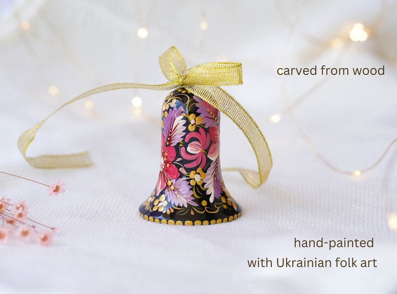 May include: A hand-painted wooden bell with a floral design in shades of pink, purple, and gold. The bell is decorated with Ukrainian folk art and is tied with a gold ribbon. "carved from wood" and "hand-painted with Ukrainian folk art" are written in the image.
