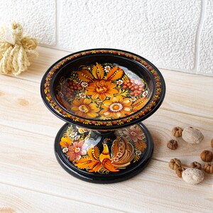 May include: A black wooden bowl with a stand, both painted with intricate floral designs in shades of orange, yellow, and red. The bowl features a bird with a long tail, while the stand has a floral pattern.