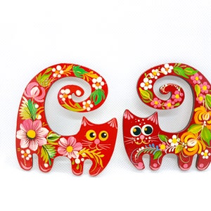 May include: Two red wooden cats with painted floral designs and curled tails. The cats have black eyes and white pupils.