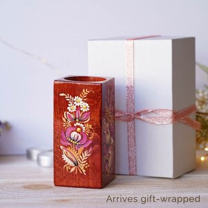 Puede incluir: Un porta l&aacute;pices de madera con un dise&ntilde;o floral pintado en la parte delantera. El porta l&aacute;pices es de forma cuadrada y est&aacute; pintado en tonos de marr&oacute;n, rosa, blanco y dorado. El porta l&aacute;pices est&aacute; sobre una superficie de madera y est&aacute; al lado de una caja de regalo blanca con una cinta rosa.