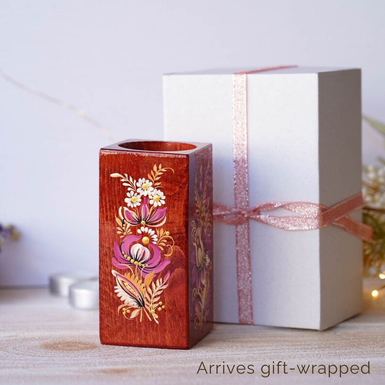Puede incluir: Un porta l&aacute;pices de madera con un dise&ntilde;o floral pintado en la parte delantera. El porta l&aacute;pices es de forma cuadrada y est&aacute; pintado en tonos de marr&oacute;n, rosa, blanco y dorado. El porta l&aacute;pices est&aacute; sobre una superficie de madera y est&aacute; al lado de una caja de regalo blanca con una cinta rosa.