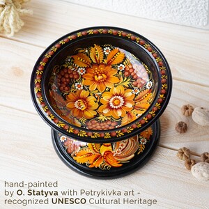 May include: A black wooden bowl with a hand-painted design of yellow and orange flowers and berries. The bowl is on a stand with a rooster design. The bowl is hand-painted with Petrykiwka art, recognized by UNESCO as a Cultural Heritage.