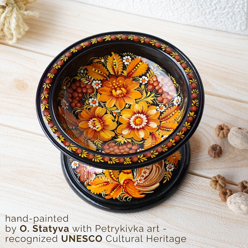 May include: A black wooden bowl with a hand-painted design of yellow and orange flowers and berries. The bowl is on a stand with a rooster design. The bowl is hand-painted with Petrykiwka art, recognized by UNESCO as a Cultural Heritage.
