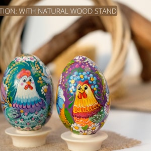May include: Two hand-painted wooden eggs with a rooster design. The eggs are on white wooden stands. The eggs are decorated with flowers and other details. The background is blurred.