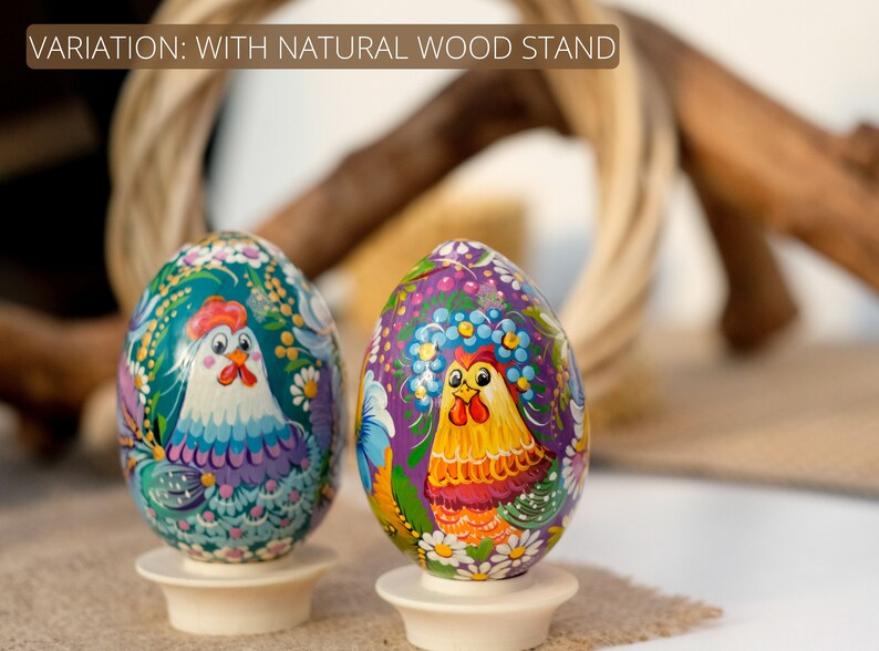 May include: Two hand-painted wooden eggs with a rooster design. The eggs are on white wooden stands. The eggs are decorated with flowers and other details. The background is blurred.