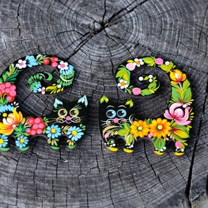 May include: Two black wooden cat figurines with colorful floral designs painted on them. The cats are curled up with their tails wrapped around their bodies.