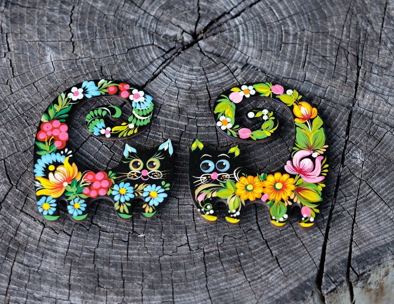 May include: Two black wooden cat figurines with colorful floral designs painted on them. The cats are curled up with their tails wrapped around their bodies.