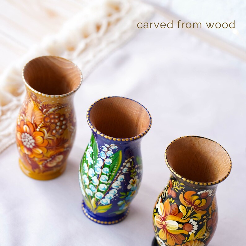 May include: Three hand-painted wooden vases with floral designs. The vases are carved from wood and have a traditional folk art style. The vases are painted in bright colors, including orange, yellow, blue, green, and black. The text "carved from wood" is visible in the image.