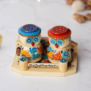 May include: A pair of wooden owl salt and pepper shakers, hand-painted with floral designs in shades of blue, orange, and yellow. The shakers are presented on a wooden tray with a blue and orange floral design.