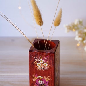 Puede incluir: Un jarr&oacute;n de madera cuadrado con una base roja pintada con dise&ntilde;os florales en rosa, blanco y amarillo. El jarr&oacute;n est&aacute; lleno de hierba de la pampa seca.