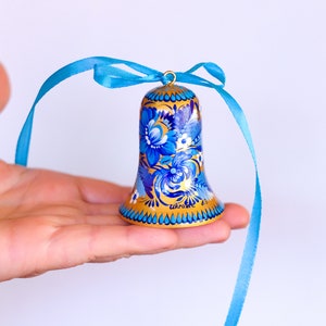 Puede incluir: Una campana dorada con dise&ntilde;os florales azules y blancos pintados en ella. La campana tiene una cinta azul atada a la parte superior.