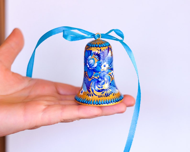 Puede incluir: Una campana dorada con dise&ntilde;os florales azules y blancos pintados en ella. La campana tiene una cinta azul atada a la parte superior.