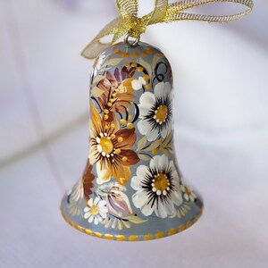 Op de afbeelding: Een grijze bel-versiering met een bloemenpatroon geschilderd in tinten bruin, wit en goud. De bel hangt aan een gouden lint.