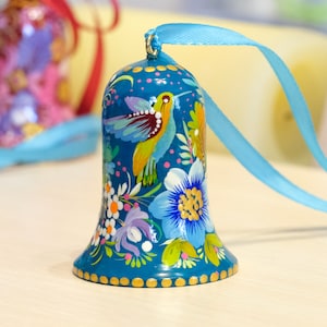 May include: A blue bell ornament with a painted design of a hummingbird and flowers. The bell has a gold trim and is hanging from a blue ribbon.