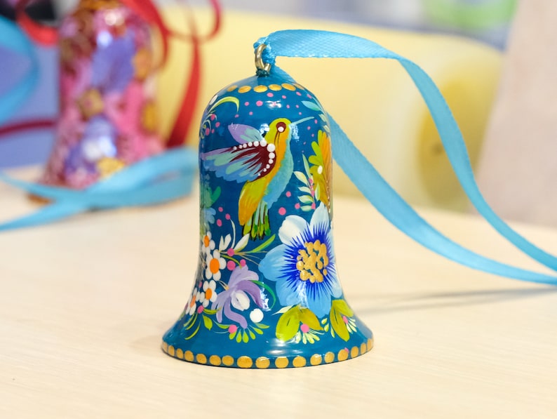 May include: A blue bell ornament with a painted design of a hummingbird and flowers. The bell has a gold trim and is hanging from a blue ribbon.